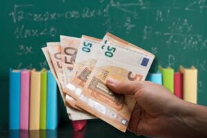 Mano con banconote da 50€ in primo piano e lavagna con libri sullo sfondo