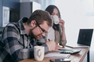 Un ragazzo che fa dei calcoli e una ragazza alle sue spalle che beve caffè